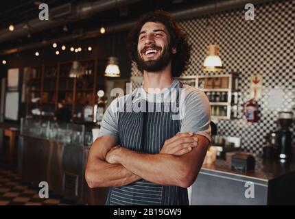 Fröhlicher junger Mann, der Schürze trägt, die mit gefalteten Armen im Kaffeehaus steht Stockfoto