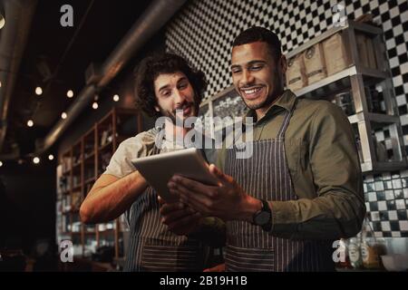 Fröhliche junge multiethnische Kollegen arbeiten im Café zusammen, indem sie ein digitales Tablet verwenden, das hinter der Theke steht Stockfoto