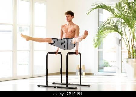 Junger Mann, der parallel Bar-Kalisthenik ausführt, übt ein V-Sit mit offenen Beinen in einer hellen High-Key-Sporthalle mit Kopierraum und vergossener Handfläche aus Stockfoto
