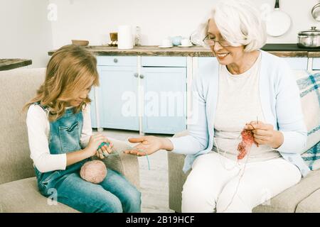 Enkelin In Der blauen Jeans Insgesamt Hilft Ihrer Grauhaarigen italienischen Granny In Legeren Kleid, Verschiedene Arten von Kleidung Zu Stricken, Während Sie Auf Sofa O Sitzt Stockfoto