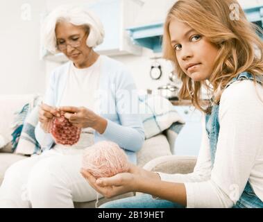 Blonde, Haarige Enkelin Hält Fäden, Die Ihrer Großmutter Helfen, Sich Zu Stricken, Während Sie An Saisonalen Feiertagen Im Wohnzimmer Im Küchenhintergrund Sitzt Stockfoto