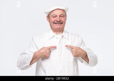 Reifer hispanischer Koch in weißer Uniform, der darauf hinweist, dass er stolz auf seinen Erfolg ist. Aufnahmen im Studio der richtigen Wahl Stockfoto