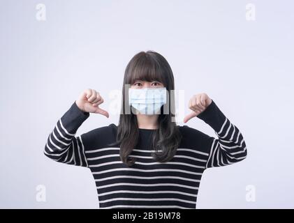 Porträt der jungen asiatischen Frau, die eine medizinische chirurgische blaue Gesichtsmaske trägt und die Daumen nach unten zeigt, die auf weißem Hintergrund isoliert ist, leer, schließen u. Stockfoto