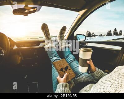 Frau trinkt Kaffeepapierbecher im Auto mit Fuß warmen Socken auf dem Armaturenbrett - Mädchen entspannt sich in Auto-Trip Lesen Reisebuch mit Schneebergen in B. Stockfoto