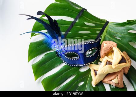 Purim, Karneval, Mardi Gras festlicher Hintergrund mit Karnevalsmaske und Dreieckscookes auf Monstera-Blatt. Purim Celebration Konzept mit Hamantaschen. Stockfoto