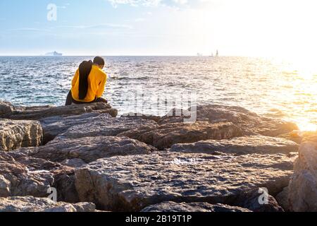Rückansicht eines Mannes in einem gelben Hemd, das auf einem Felsen am Meer sitzt. Freizeit und Entspannungskonzept. Stockfoto