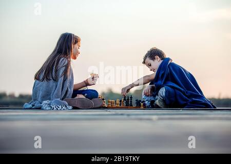 Zwei kleine, niedliche kleine Freunde, Junge und Mädchen, die am Abend beim Schachspielen am See Spaß haben. Kinder spielen. Freundschaft. Stockfoto