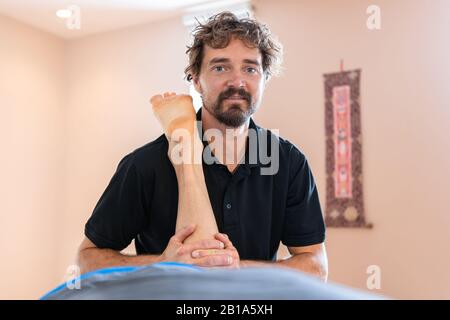 Ein geschweifter Sportmassage-Therapeut, der die Kamera betrachtet. Professionelle Massageanwendungen, die einem Sportler Massageverfahren geben Stockfoto