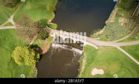 Luftdrone Blick auf eine malerische Straßenbrücke über einen englischen Fluss, der über ein kleines Wehr führt. Stockfoto