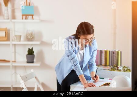 Professionelle Designerin genießt ihre Arbeit im Büro mit modernem Interieur, modiert das Modell des Gebäudes im Hintergrund des Fotos. Stockfoto