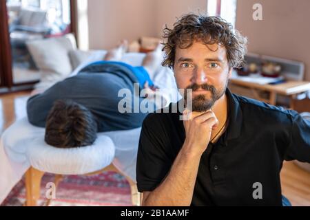 Portrait für Sportmassage-Therapeuten. Ein professioneller Masseur, der in der medizinischen Klinik posiert und im Hintergrund einen Patienten auf dem Massagetisch liegt Stockfoto