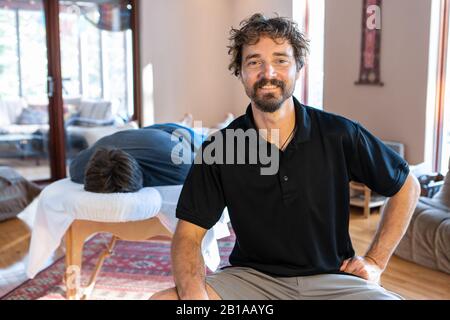 Porträt des lächelnden Physiotherapeuten am Arbeitsplatz. Ein starker männlicher Klient, der auf dem Tisch liegt und auf die Behandlung der Körpermassage wartet Stockfoto
