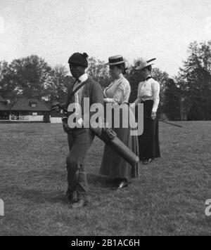 Golf: Afroamerikanischer Caddy und 2 weiße Golferinnen stehen und warten auf einen weiteren Golfer. Caddy hat eine dünne Golftasche, die von seiner Schulter hängt. Das Clubhaus ist im Hintergrund. Foto ca. 1910 um meine anderen Vintage-Bilder im Zusammenhang mit dem Golf zu sehen, Suche: Prestor vintage Sport African Stockfoto