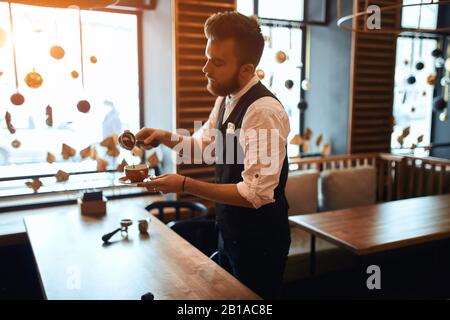 Bärtiger talentierter Barista, der Kaffee in die Tasse schüttet, während er im Café neben dem Tisch steht, Nahaufnahme der Seitenansicht Stockfoto