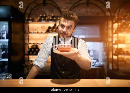 Lächelnder attraktiver Mann, der seinen Kunden im Coffeeshop eine Tasse Tee anbietet: Lifestyle, Freizeit, Interesse Stockfoto