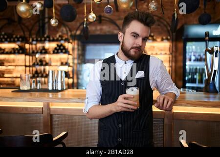 Gutaussehender junger bärtiger Mann, der die Kaffeetasse hält und die Kamera ansieht, während er an der Theke steht, Nahaufnahme Stockfoto
