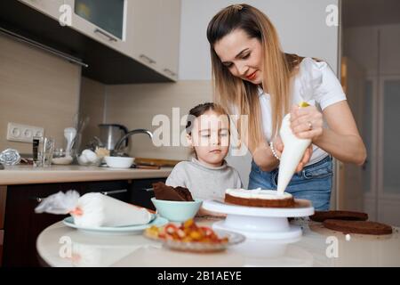 Junge attraktive Frau, die Sahne auf die Kuchenoberseite legt, kleine neugierige niedliche Mädchen, die es beobachtet, Nahaufnahme Foto: Familientradition, Freizeit, f Stockfoto