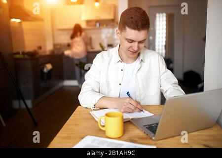 Der junge glückliche Mann freute sich über seinen erfolgreichen Deal und machte sich beim Frühstück Notizen, die Frau machte Hausaufgaben im verschwommenen Hintergrund, Nahaufnahme Stockfoto