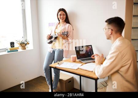 Junge schöne fröhliche brünette Frau, die sich an die Wand lehnt und Lieder für ihren Freund singt, der am Tisch sitzt und einen Laptop verwendet. Nahaufnahme Stockfoto