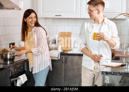 Junges fröhliches Positivpaar, das das Rezept des Gerichts bei der Arbeit in der Küche diskutiert. Seitenansicht Foto. Mann mit einem gelben Becher und einem Gespräch w Stockfoto