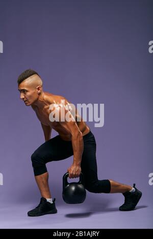 Seitenansicht in voller Länge, bei der der junge Mann ein Knie knickt, das eine Kettleblocke in den Händen hält, isolierter blauer Hintergrund, Studioaufnahme, exellentes Trainingsprogramm, Stockfoto