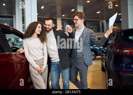 Bärtiger gutaussehender Mann, der den Schlüssel für ein neues Luxusauto im Autohaus gibt, fröhlicher Ladenassistent, der neuen Besitzern eines modernen Autos bei Dealer Cent gratulierte Stockfoto