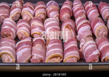 Rohe Würstchen in Speck (manchmal Schweine in einer Decke genannt), in Reihen in einem Backblech, bereit zum Ofengebacken. Stockfoto