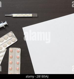 Vorlage für weißes Papier mit Pillen in Blister, Thermometer und Spritze zur Injektion auf dunklem, wengem Holzhintergrund. Die Bereitstellung medizinischer und pharmakologischer Dienstleistungen. Stockfoto