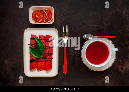 Nahaufnahme von Köstlichen hausgemachten roten Orangen dünne Pfannkuchen mit frischen Orangenscheiben und rotem Fruchttee. Draufsicht, Nahaufnahme auf dunklem Hintergrund Stockfoto