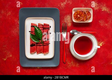Nahaufnahme von Köstlichen hausgemachten roten Orangen dünne Pfannkuchen mit frischen Orangenscheiben und rotem Fruchttee. Draufsicht, Nahaufnahme auf rotem Hintergrund Stockfoto