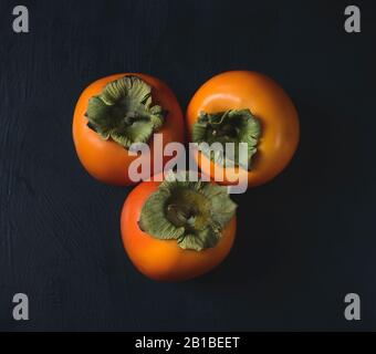 Хурма. Persifonen auf dunklem Hintergrund. Persifonen auf einem Holztisch. Drei reife Persifonen auf dunklem Hintergrund. Stockfoto