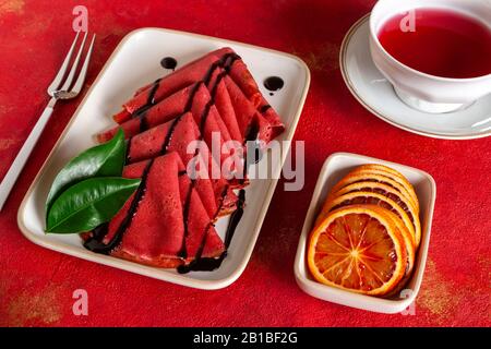 Nahaufnahme von Köstlichen hausgemachten roten Orangen dünne Pfannkuchen mit frischen Orangenscheiben und rotem Fruchttee. Draufsicht, Nahaufnahme auf rotem Hintergrund Stockfoto