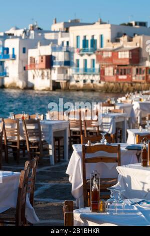 Helle, malerische Nachmittagssicht auf einen leeren Tisch, der auf einen Sonnenuntergang wartet, der am Hafen in der griechischen Altstadt von Mykonos eingerichtet wurde Stockfoto