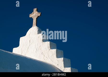 Steinernes Kreuz, das auf rauem weißem Stuck-Kirchturm der traditionellen griechischen Inselkirche unter hellblauem mediterranen Himmel steht Stockfoto