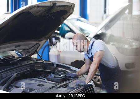 Portrait des muskulösen Automechanikers, der bei der Inspektion im Garagengeschäft in die offene Haube des Fahrzeugs blickt, Kopierraum Stockfoto