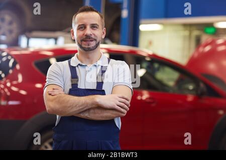 Taille oben Porträt des muskulösen Automechanikers stehend mit den Armen, die gegen rotes Auto gekreuzt werden, während sie in der Kfz-Werkstatt posieren, Kopierraum Stockfoto