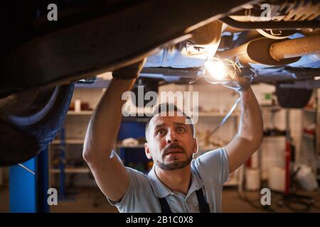 Hochwinkelansicht bei einem reifen Mechaniker, der bei der Inspektion in der Werkstatt für automatische Reparaturen unter dem Fahrzeug auf dem Aufzug betrachtet und die Lampe hält, Kopieraum Stockfoto