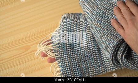 Eine Person zeigt einen handgewebten Wollschal aus Webstühlen. Gewebtes Material mit weißen Streifen und dunkleren Bändern in blau-grünen Tönen in Strickhänden. Stockfoto