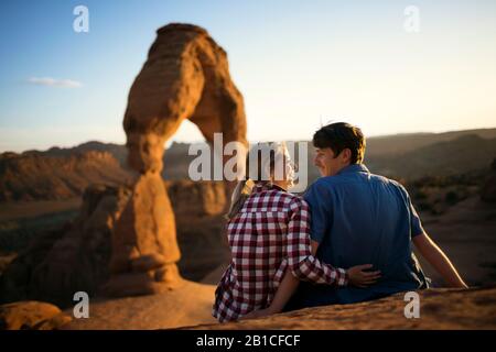 Junge Paare, die sich gegenseitig betrachten, während sie auf einer Felsformation sitzen, die einen malerischen Canyon überblickt Stockfoto