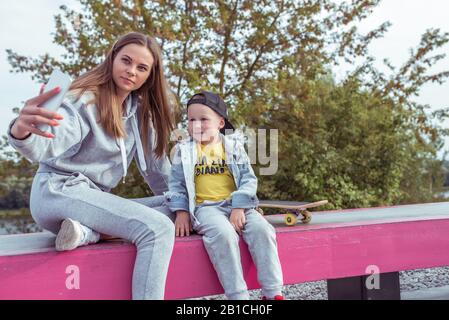 Frau Mama macht Fotos am Telefon, selfie, kleiner Junge, Sohn 3-5 Jahre alt, entspannen mit Skateboard, Sommer auf Sportplatz, Emotionen Glück, Spaß Stockfoto