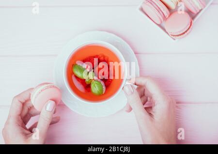 Weibliche Hände, die eine weiße Tasse Himbeertee und einen pinkfarbenen Makronen auf einem blassrosa Hintergrund halten. Draufsicht Stockfoto