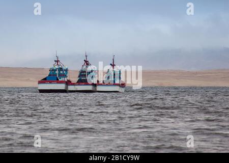 Drei identische Fischerboote, die in einer Linie vermocht sind, betonen die Gegennatur des Ozeans und der Wüste, während sie sich in Paracas treffen. Stockfoto