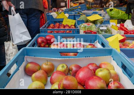 Äpfel auf blauem Plastikkorb vor dem Stall außerhalb des Lebensmittelgeschäftes neben dem Flur des Freiluftmarktes mit unscharfem Hintergrund von Menschen. Stockfoto