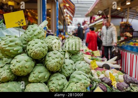 Haufen Artischockengemüse vor dem Festplatz außerhalb des Lebensmittelgeschäftes neben dem Korridor des Freiluftmarktes mit unscharfem Hintergrund von Menschen. Stockfoto