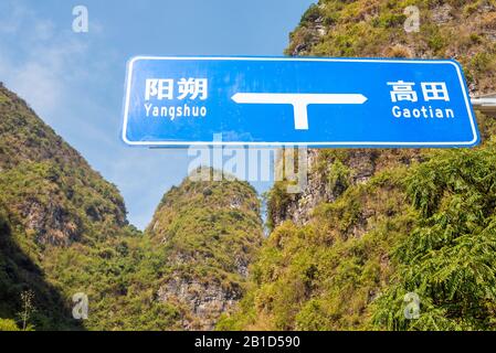 Blaues Straßenschild mit Yangshuo und Gaotian, das sowohl in chinesischer als auch rumänischer Schrift gegen Karsthügel und Vegetation an einem sonnigen Tag in Yangshuo, Provinz Guangxi, China, geschrieben wurde Stockfoto