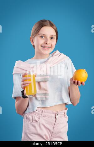 Porträt eines lächelnden, süßen Mädchens, das vor blauem Hintergrund steht und ein Glas Orangensaft hält Stockfoto