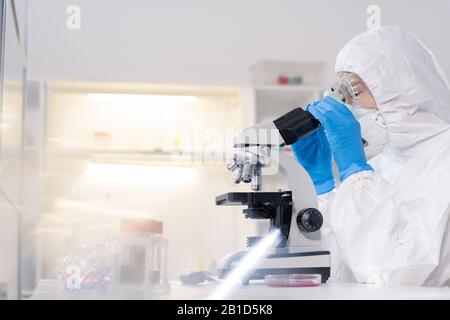 Fokussierter Labortechniker in Schutzanzug und Maske mit Mikroskop bei der Arbeit mit Blutproben Stockfoto