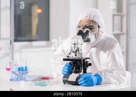 Forschungswissenschaftler der konzentrierten asiatischen Virologie in Schutzanzug, Maske und Atemschutzmaske mit Mikroskop bei der Analyse von Proben im Labor Stockfoto