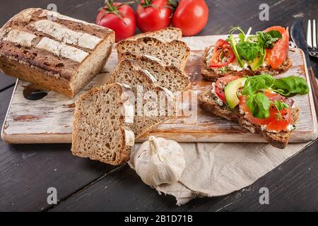 Leckeres hausgemachtes Brot, geröstete Sandwiches mit Räucherlachs, Avocado, Tomaten, Kräutern und Käse auf einem Holzschneidebrett Stockfoto