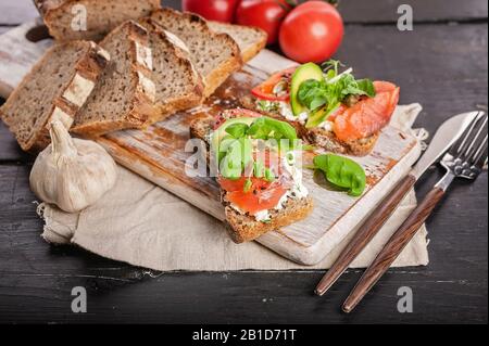 Hausgemachtes Brot mit getoasteten Sandwiches mit Räucherlachs, Avocado, Tomaten, Kräutern und Käse auf einem Holzschneidebrett Stockfoto